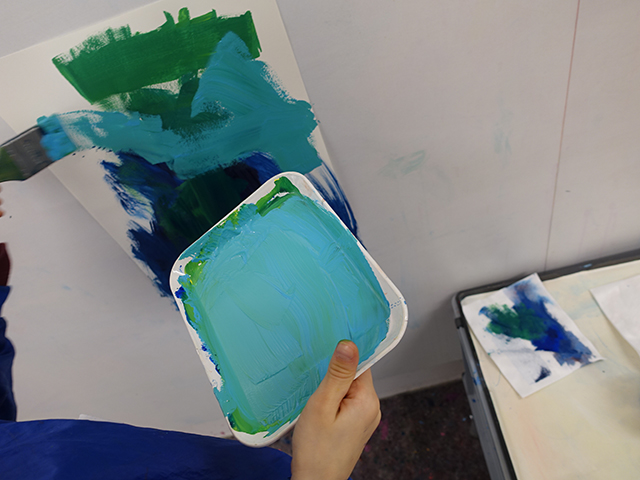 Kind mit Palette in Hand, malt ein Bild mit Türkis, Dunkelblau und Grün.